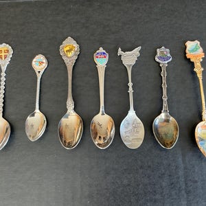 May include: A selection of souvenir spoons with decorative handles and enamel crests. The spoons are silver-toned, featuring various designs including landmarks and animals. The crests are detailed and colourful. The spoons vary in length, with the longest being approximately 15 cm.