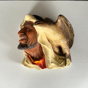 Könnte beinhalten: Eine detaillierte Skulptur eines Mannes mit dunkler Hautfarbe, der einen Turban trägt, mit einem Falken auf seiner Schulter. Die Skulptur zeigt einen cremefarbenen Turban, ein rot-gelbes Gewand und einen Falken mit braunen und beigen Federn.