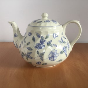 Puede incluir: Una tetera de cerámica blanca con un patrón floral azul. La tetera tiene una tapa y un asa.