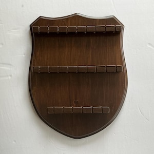 May include: A wooden spoon display rack in a shield shape. The rack is stained a dark brown color and has three rows of small rectangular slots to hold spoons. The rack is designed to be mounted on a wall.