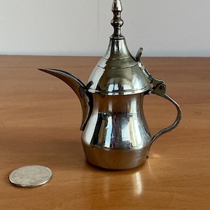 May include: A small, silver-coloured metal coffee pot with a curved spout, handle, and ornate lid. A coin is next to the pot for scale. The pot has a reflective surface.