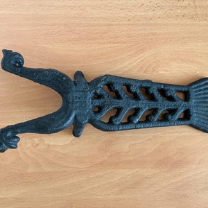 May include: A black cast iron boot jack featuring a bifurcated end with two curled tips and a central section with a decorative, openwork fishbone pattern that terminates in a flat, rectangular base with vertical ridges for stability.