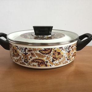 Peut inclure: Une casserole en émail blanc avec une poignée et un couvercle noirs. La casserole est décorée d'un motif floral brun et jaune.
