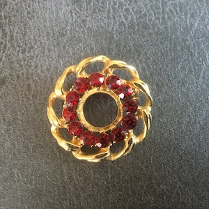 May include: A gold-toned brooch with a circular design featuring a ring of red rhinestones.