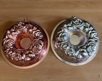 Puede incluir: Dos moldes decorativos para bizcocho. Uno es de color cobre, el otro plateado. Ambos tienen forma circular con un agujero central y están decorados con un patrón de rosas. Cada molde tiene un pequeño lazo para colgar.