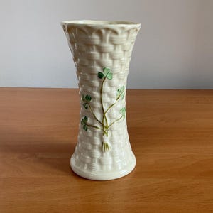 Könnte beinhalten: Eine cremefarbene Keramikvase mit einem strukturierten Korbgeflechtmuster auf der Außenseite. Die Vase zeigt ein handgemaltes grünes Kleeblatt-Design auf der Vorderseite, mit einem ausgestellten Rand und einem runden Boden, aufrecht stehend.