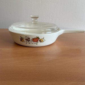 Może przedstawiać: Biała ceramiczna patelnia z długą rączką i przezroczystą szklaną pokrywką. Zewnętrzna strona patelni ozdobiona jest kolorowym nadrukiem przedstawiającym czerwonego pomidora, brązowe grzyby i zielone zioła ułożone w poziomym rzędzie.