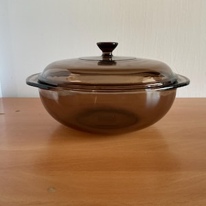 Puede incluir: Una fuente de vidrio marrón con tapa a juego. El plato tiene forma de cuenco redondeado con un pomo marrón oscuro en la parte superior de la tapa. El plato está sobre una superficie de madera.