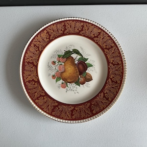 Puede incluir: Plato decorativo redondo con centro blanco y borde granate con diseño de cachemira dorada. El centro presenta una pera, una ciruela, frambuesas y nueces. El plato tiene un borde dorado.