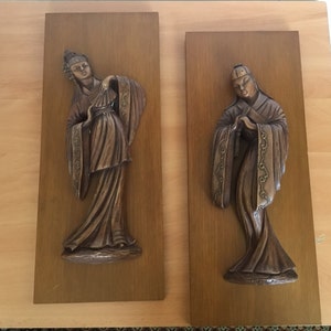 Op de afbeelding: Twee houten wandplaquettes met reliëfsculturen van figuren in traditionele Aziatische kleding. De figuren zijn gesneden in een donkerbruine kleur en hebben ingewikkelde details.