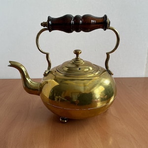 Könnte beinhalten: Eine Messing-Teekanne mit einem Holzgriff und einem Deckel. Die Teekanne hat einen runden Körper und einen Ausguss. Die Teekanne steht auf einer Holzoberfläche.