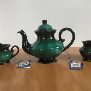 Puede incluir: Un juego de tetera, jarra de crema y azucarero de cerámica verde y negro. La tetera tiene una tapa y un asa negros. La jarra de crema y el azucarero tienen bordes negros.