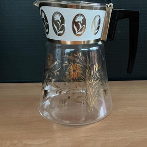 Puede incluir: Una cafetera de vidrio vintage con una banda blanca con diseños florales dorados. La cafetera tiene un cuerpo de vidrio transparente con patrones dorados de trigo y flores, un asa negra y un borde dorado.