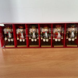 May include: A set of six wooden nutcracker place card holders in a red display box. Each nutcracker features a painted uniform in red, green, or brown, a gold crown or hat, a white faux fur beard, and a square base for holding a place card.