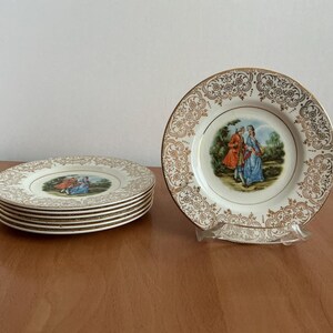 May include: Set of vintage decorative plates with gold filigree rims and a central scene of a couple in period clothing. One plate is displayed upright, while the others are stacked. The plates are on a wooden surface.