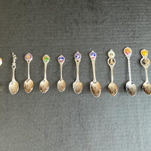 May include: A selection of souvenir spoons with ornate handles. The spoons are crafted from silver-coloured metal and showcase diverse designs, such as enamel crests, figurines, and landmarks. The spoons are displayed in a line on a dark background.