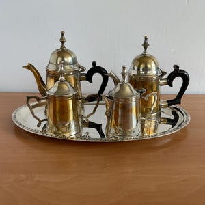Peut inclure: Un service à thé argenté composé de deux théières, d'un sucrier et d'un pot à lait, tous dotés de poignées et de boutons noirs. Les quatre pièces sont disposées sur un plateau de service ovale argenté avec un bord perlé décoratif.