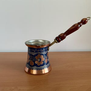 Puede incluir: Una cafetera de cobre con borde acampanado y un mango largo de madera marrón oscuro. El cuerpo presenta un acabado pintado en azul profundo decorado con intrincados motivos florales dorados y un borde blanco en forma de rejilla.