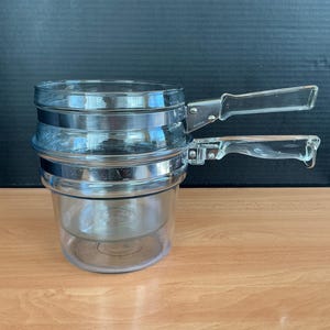 Op de afbeelding: Een heldere glazen stoompan met twee heldere glazen handvatten. De pot heeft drie secties met zilveren metalen banden. De pot staat op een lichtbruin houten oppervlak.