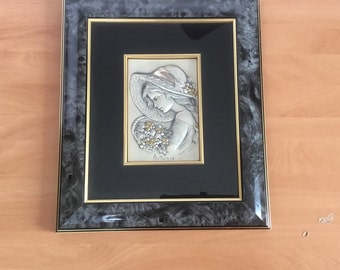 May include: A framed silver relief sculpture of a woman in a hat holding a basket of grapes. The sculpture is set against a black background and the frame is black with a gold trim. The text "Autunno" is visible at the bottom of the sculpture.