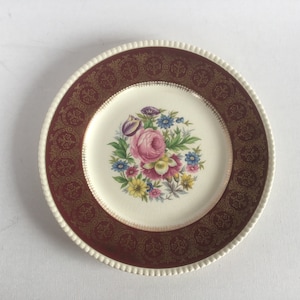 Puede incluir: Un plato de cena de cerámica blanca con un borde burdeos y dorado. El centro del plato presenta un diseño floral con una rosa rosa y otras flores de colores.