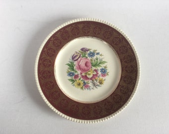 May include: A white ceramic dinner plate with a burgundy and gold border. The center of the plate features a floral design with a pink rose and other colorful flowers.