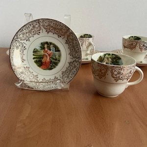 Puede incluir: Un juego de té vintage con tazas y platillos blancos con filigrana dorada y una imagen central de una mujer con un vestido rojo. El juego incluye cuatro tazas y platillos, ideal para servir té. Las tazas y platillos son de porcelana.