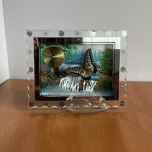 May include: A clear glass frame with a silver-coloured border and a snowflake design. Inside the frame is a painting of a waterfall with a butterfly flying over it. A small clock is also visible in the painting.