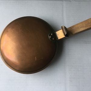 Peut inclure: Une poêle en cuivre et en bois avec une poignée en bois. La poêle est ronde et a une surface lisse. La poignée est fixée à la poêle par une charnière en métal.