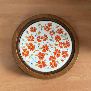 Op de afbeelding: Ronde houten onderzetter met een wit keramisch middenstuk. Het keramiek heeft een bloemenpatroon van oranje bloemen, groene bladeren en blauwe accenten. De onderzetter is ontworpen om oppervlakken tegen hitte te beschermen.