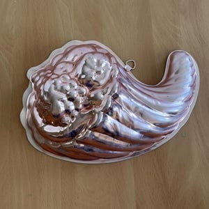 Pode incluir: Uma forma de metal vintage de cor cobre em forma de cornucopia. A forma tem uma superfície lisa e brilhante e é projetada para assar.