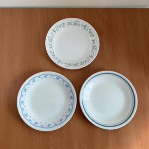 May include: Three white ceramic plates with different blue floral designs. One plate has a delicate floral and leaf pattern around the rim. Another has a blue floral pattern, and the third has two blue stripes around the rim. A set of dinnerware.