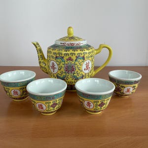 May include: A yellow ceramic tea set featuring a teapot and four teacups. The teapot and cups have a decorative floral and Chinese character design. The set is on a wooden surface.