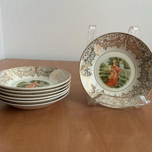 May include: Set of vintage decorative plates. Each plate features a central image of a woman in a red dress in a landscape scene. The plates have a gold filigree border and are displayed on a wooden surface.