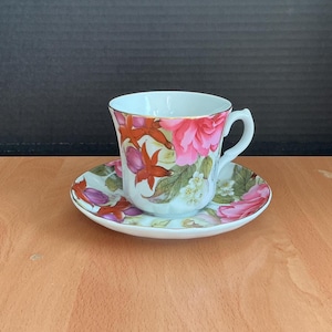 May include: A white teacup and saucer set with a floral design featuring pink roses, red leaves, and white blossoms. The cup has a gold rim and a curved handle. The saucer mirrors the cup's floral pattern.