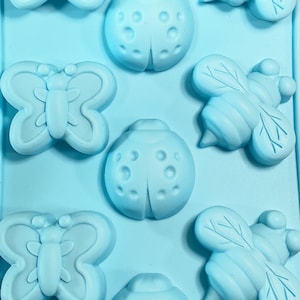 May include: Blue silicone mold with six cavities shaped like butterflies, ladybugs, and bees. The mold is designed for making small cakes, candies, or other treats.