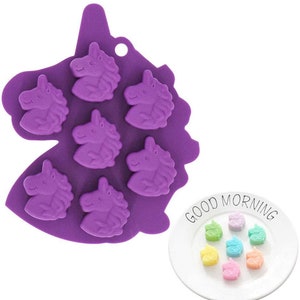 May include: A purple silicone mold shaped like a unicorn head, with seven unicorn face cavities. The mold is next to a white plate with six small, colorful unicorn-shaped candies. The plate has the words "GOOD MORNING" printed on it.