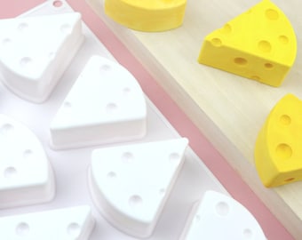 Könnte beinhalten: Eine weiße Silikonform mit acht keilförmigen Vertiefungen, jede mit einem Muster aus kleinen Kreisen, die an Käse-Löcher erinnern. Es gibt drei gelbe käseförmige Objekte auf einer Holzoberfläche.