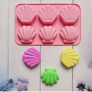 May include: A pink silicone seashell mold with six shell-shaped cavities. Three colorful seashell-shaped soaps are also present: pink, yellow, and green. The mold and soaps are on a white wooden surface.