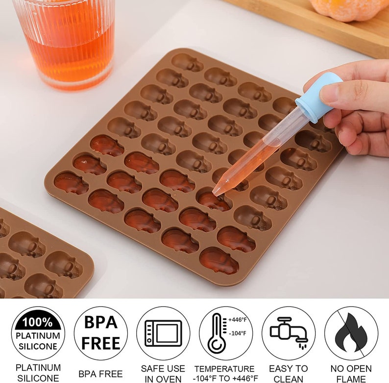 May include: Brown silicone mold with 48 skull-shaped cavities filled with a clear, orange liquid. The mold is on a white surface. The text on the image reads: "100% Platinum Silicone", "BPA Free", "Safe Use in Oven", "Temperature -104&deg;F to +446&deg;F", "Easy to Clean", and "No Open Flame".