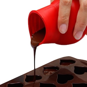 May include: Red silicone chocolate mold with a heart shape. A red silicone pouring pitcher is pouring melted chocolate into the mold.