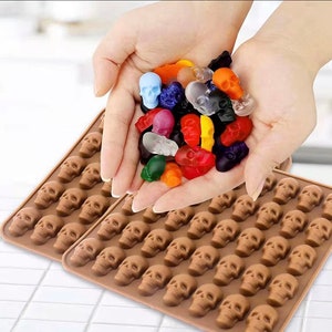 May include: Two brown silicone molds with skull shapes. A hand is holding a variety of colorful candy skulls.