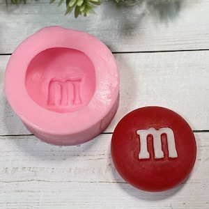May include: Pink silicone mould with an imprint of the letter "M" and a red sweet with a white "M" on top.