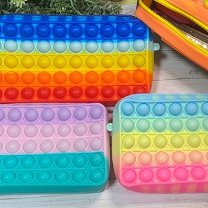 May include: Four colorful zippered pouches with a pop-it design. The pouches are rainbow colored with blue, yellow, orange, red, purple, green, and pink.