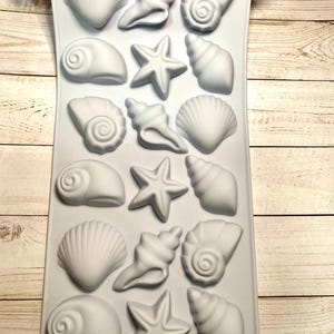 May include: A light gray silicone mold featuring various seashell and starfish shapes. The mold includes detailed depictions of conch shells, spiral shells, and starfish, perfect for crafting coastal-themed items. The mold is set against a light wood grain background.