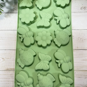 May include: Green silicone mold with 12 cavities shaped like butterflies, ladybugs, and flowers. The mold is on a white wooden surface.