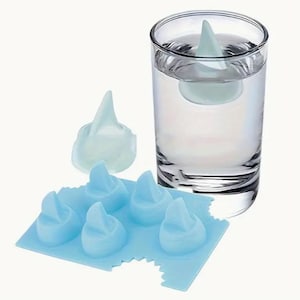 May include: A blue silicone ice cube tray with six shark fin shaped ice molds. One shark fin ice cube is floating in a glass of water.
