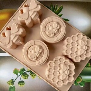 May include: A silicone mold for making soap or candles. The mold has six cavities, each with a different design. The designs include a bee, a honeycomb, and the words "Bee Honey".