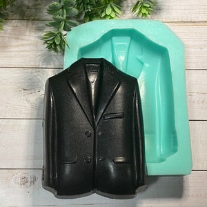May include: A black soap shaped like a blazer with two buttons, next to a light blue silicone mold in the shape of a blazer. The soap has a shiny finish and is set against a light wood background.
