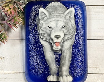 May include: A rectangular soap bar featuring a detailed wolf design. The wolf is rendered in gray and white, set against a textured, deep blue backdrop. The soap sits on a white wooden surface, with greenery visible in the background.